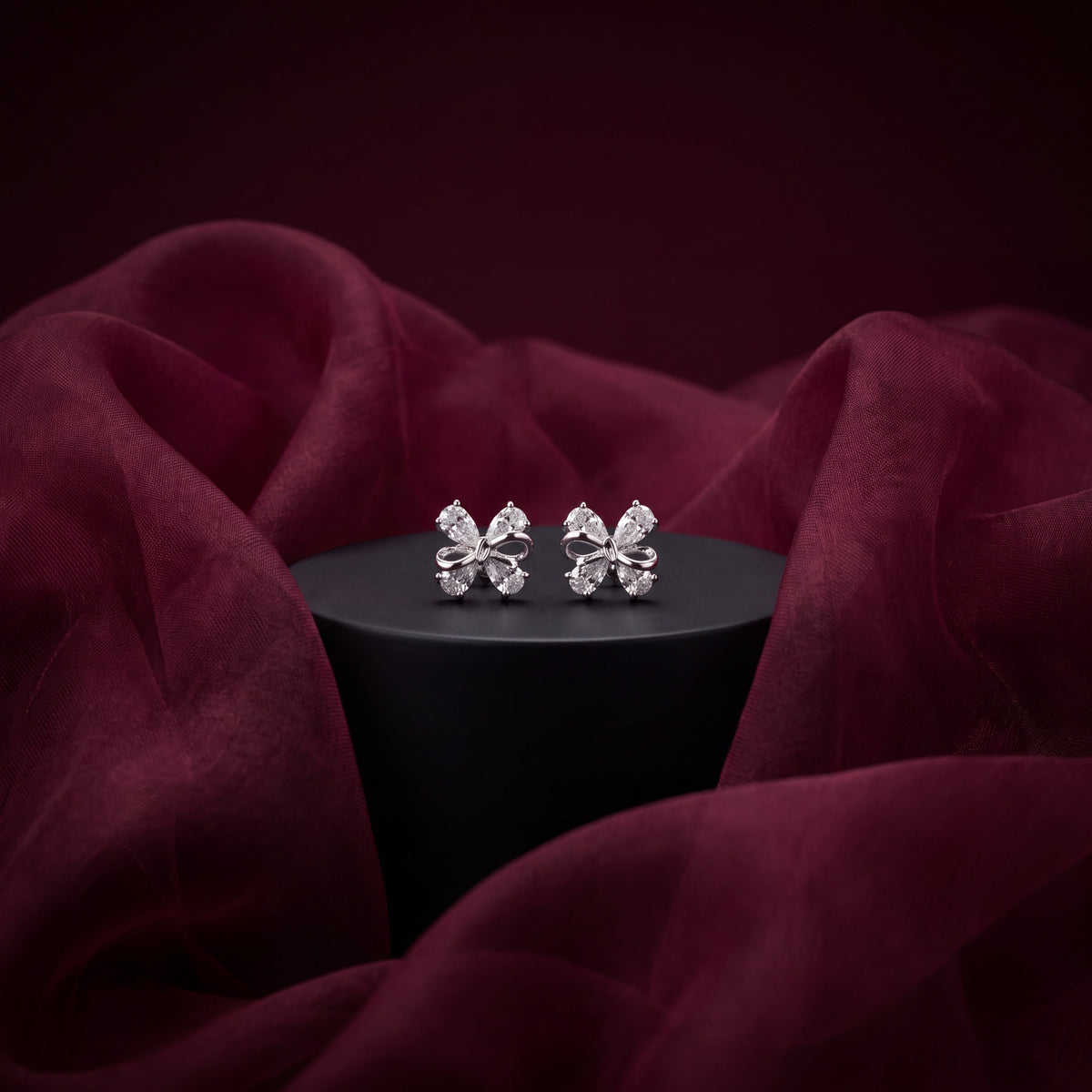 CZ Bow Stud Earrings - Silver tone on black pedestal with burgundy fabric drape lifestyle shot