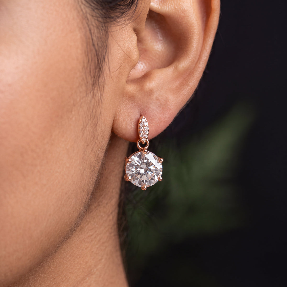 CZ Solitaire Drop Huggie Earrings - Rose Gold tone worn on model ear with dark background close up