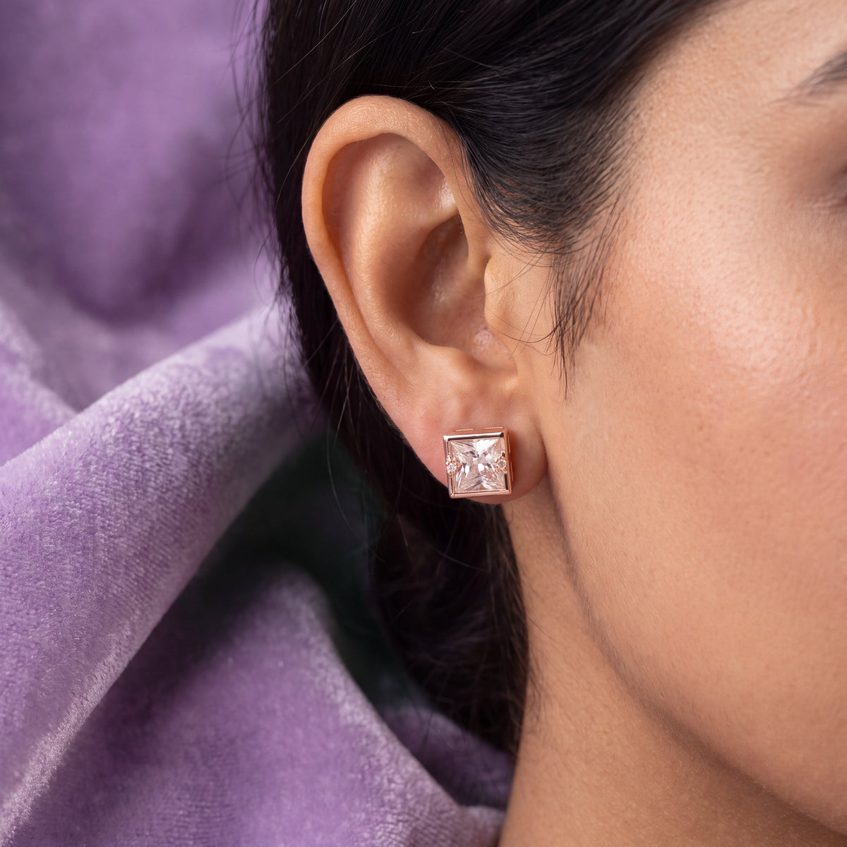 Princess Cut CZ Square Bezel Stud Earrings - Rose Gold tone worn on model ear with lilac velvet drape background close up